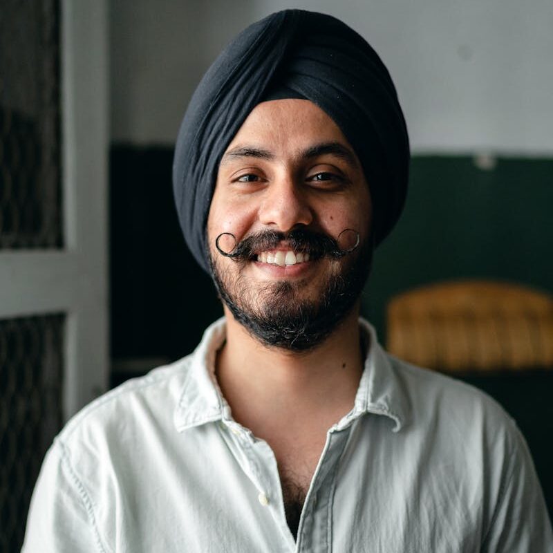 Positive bearded Indian male in shirt and turban standing in room and smiling while looking at camera