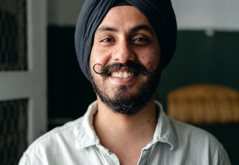 Positive bearded Indian male in shirt and turban standing in room and smiling while looking at camera